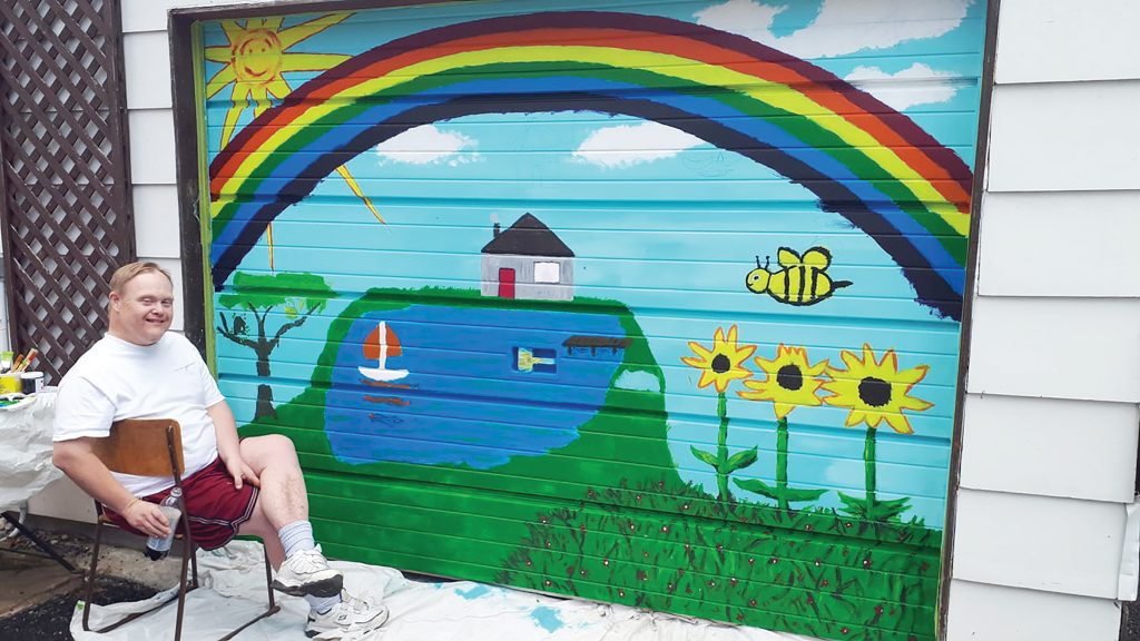 A photo of Paul Knoll sitting in front of a garage door mural in Westboro.