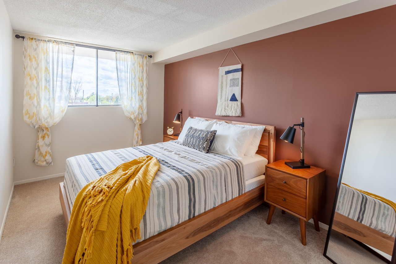 A bedroom is seen with a queen bed with a yellow blanket on the edge of it and a rose coloured wall behind it.