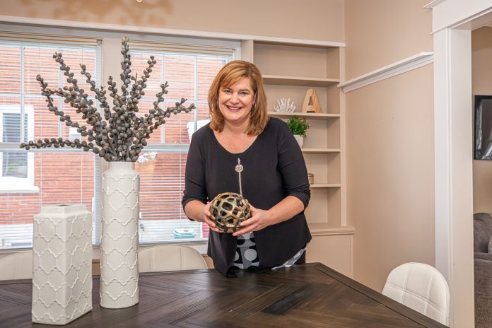 A woman stands in a living room next to a dining room table and holds a centrepiece of a globe.|