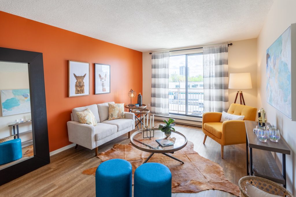 A living room with a white couch, yellow chair, blue ottomans and an orange wall is seen with a large set of windows