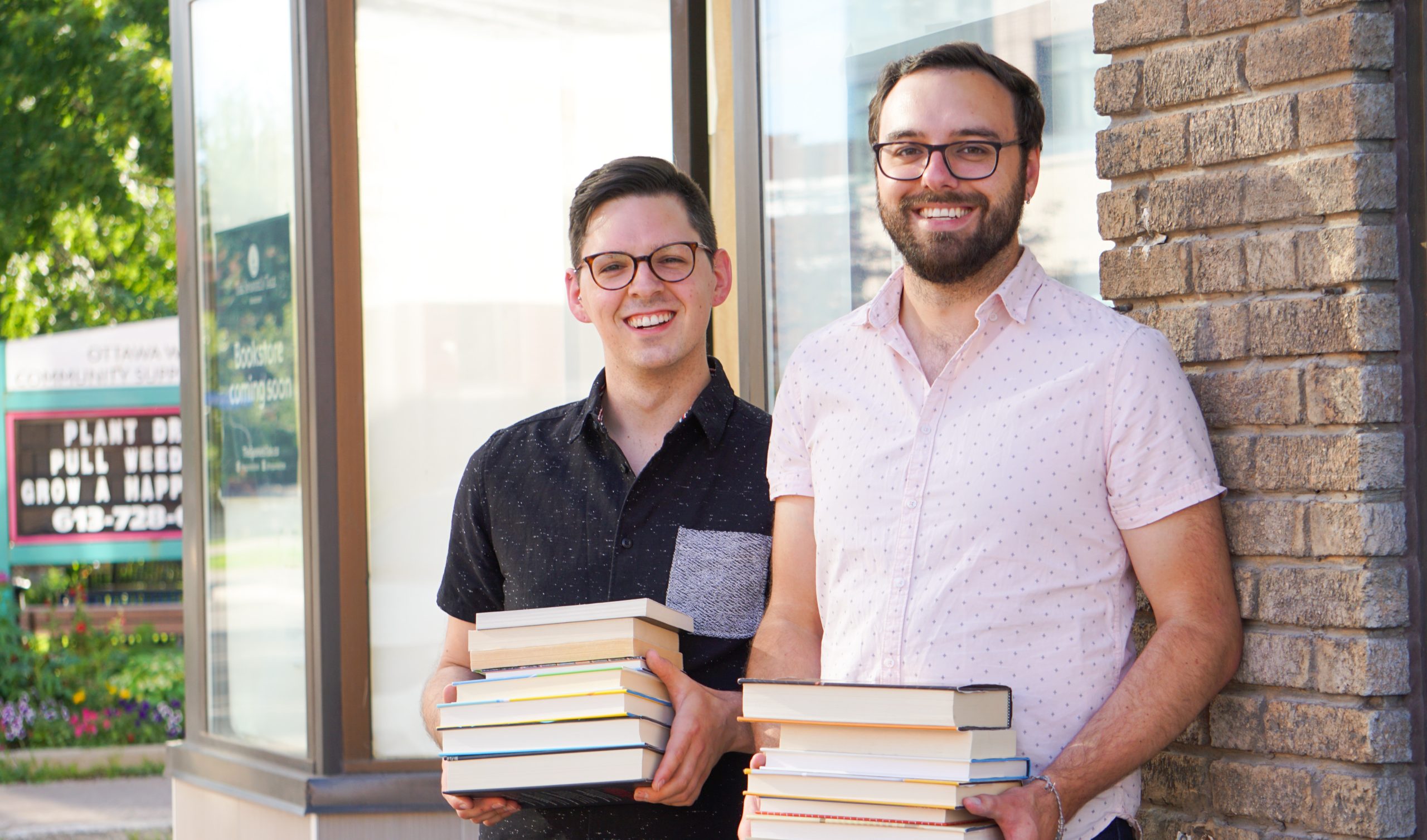 Two men stand side by side holding piles of books outside of The Spaniel's Tale bookstore.