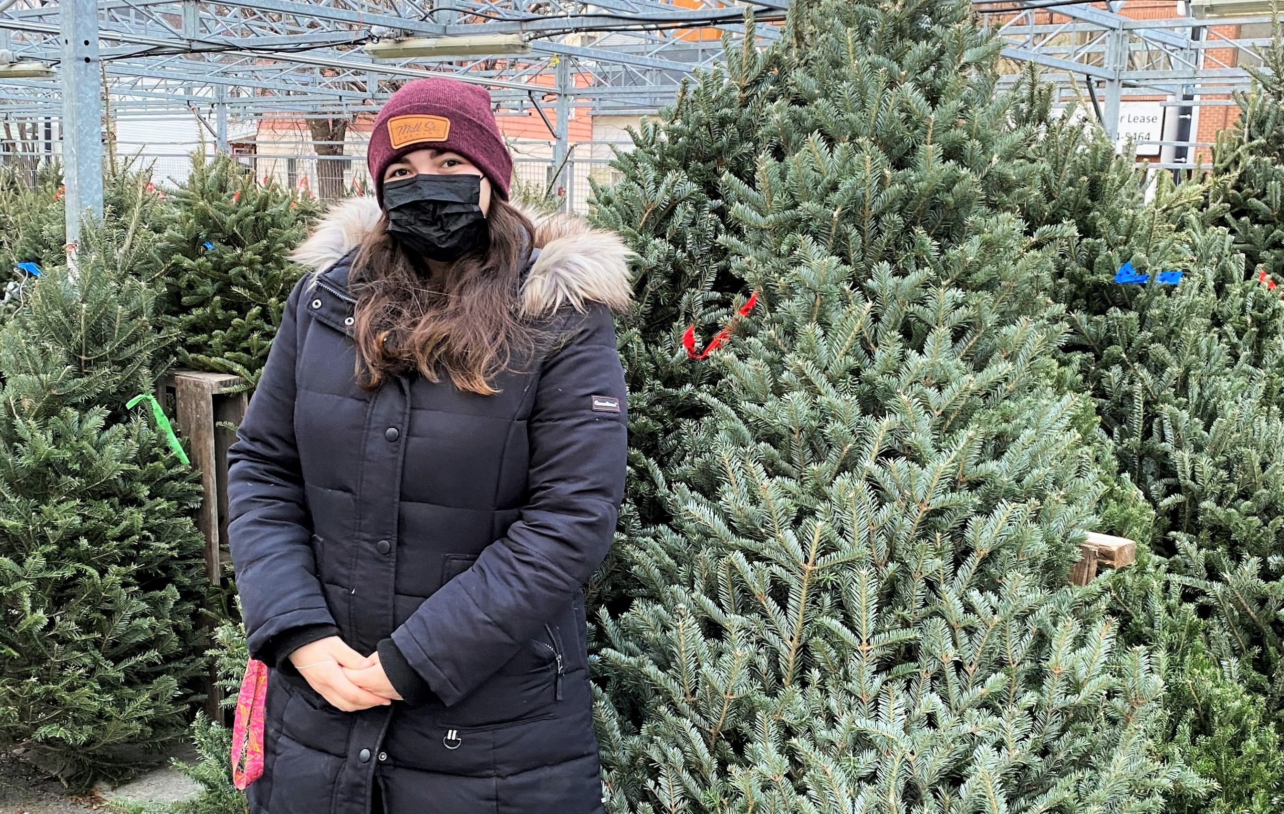 A woman wearing a black coat and black mask stands in front of Christmas trees at Parkdale Market