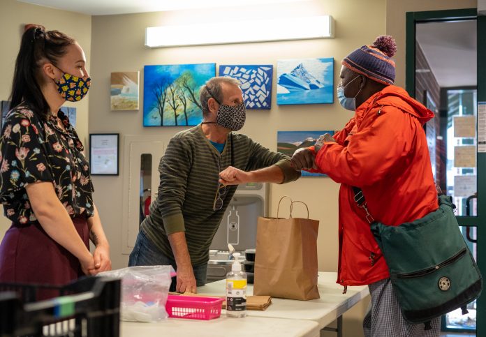 Two men bump elbows while wearing masks at a table with a woman wearing a mask watching.|