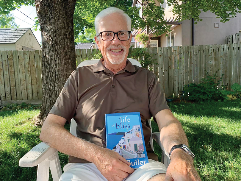 A man sits in a chair in a yard in Ottawa holding a book called "A Life of Bliss." There is a wooden fence and tree behind him and it is a sunny day. 