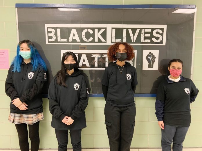 Four student stand in a row wearing black in front of a Black History month posterboard