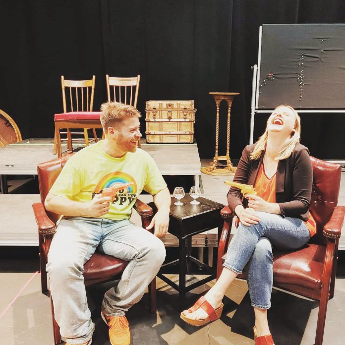 A man and a woman sit side by side in leather arm chairs on a stage. They both have fake guns pointed at one another and they are laughing.