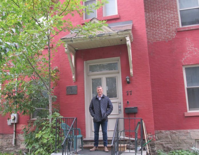 A man wearing a black jacket and blue jeans stands in front of a red brick building in Centretown.