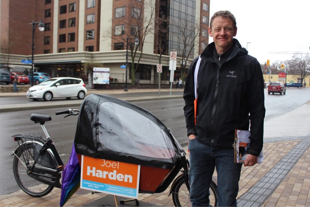 Joel Harden stands on the street next to a campaign sign on a bike in Ottawa