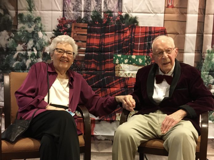 Two people sit in chairs at a retirement community with red plaid and holiday decor behind them.|