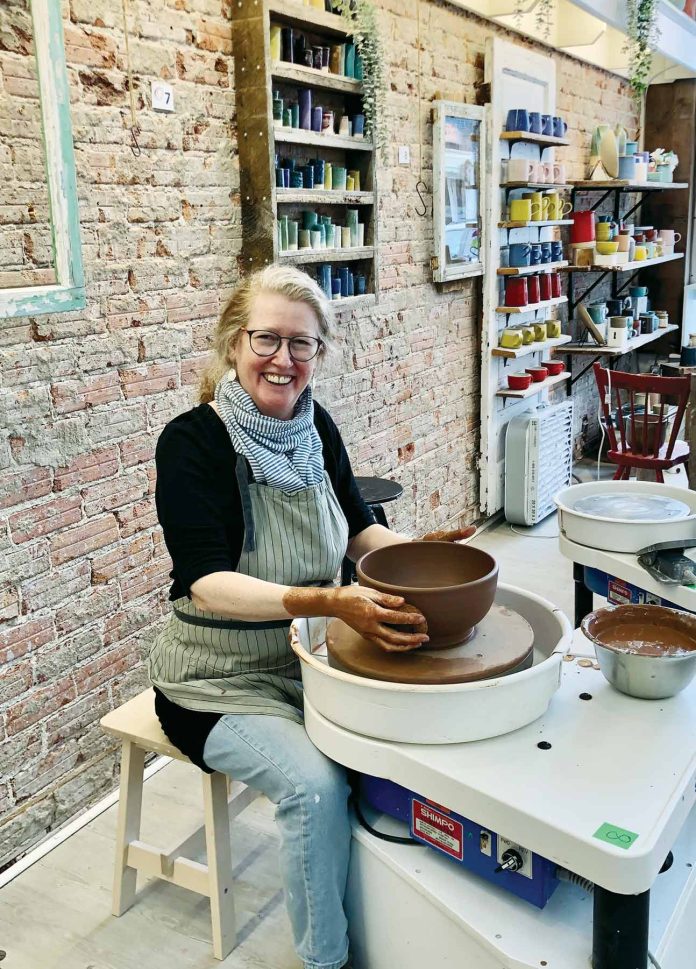 A woman sits next to a pottery wheel with a bowl of clay in front of her.|