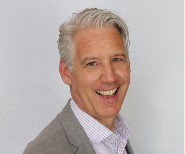 A headshot of a man against a grey background. He is wearing a light grey suit.|