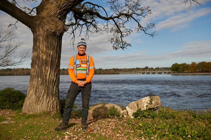 A man stands next to a tree in front of the Ottawa River on a sunny day||