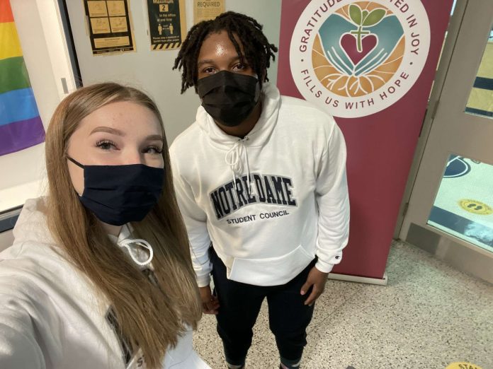 A female and male student stand side-by-side at Notre Dame High School and take a selfie photo on a phone.|