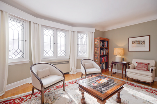 A living room of an historic home is pictured with vintage white chairs and a wood table and bookshelf|