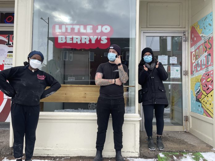 Three people in masks stand outside of Little Jo Berry's in Kitchissippi. The staff are all wearing black.||A woman places a colourful box on a table next to other colourful boxes inside Little Jo Berry's bakery in Ottawa.|A bunch of brown cupcakes with white icing and pink sparkles are shown. The cupcakes have white hearts with phrases written on them.