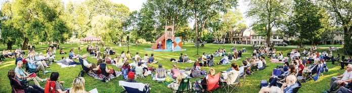 A wide shot of a crowd sitting in lawn chairs and on blankets watching a Shakespeare in the Park performance outside on a sunny day in an Ottawa park.|A cast of actors stand on the blue and orange multilevel set for Shakespeare in the Park. There is a forest behind them.|A Black actor wearing a jacket with orange ribbons flowing off off of it performs in the park.|Three actors carry a white and red boat in costume in a play in the park.|