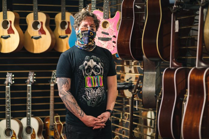 A man stands in the middle of a music store surrounded by guitars on the walls. He is wearing a t-shirt with a skull on it and wearing a multi-coloured mask.|