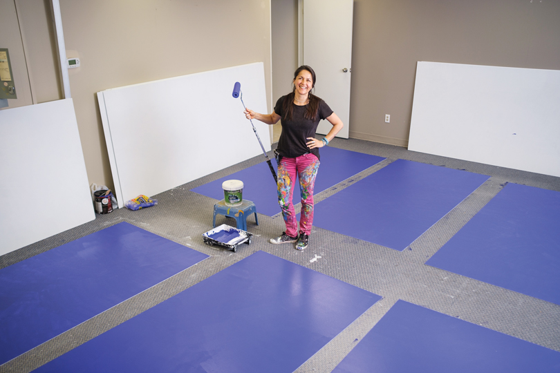 Claudia Salguero stands with a paint roller and blue panels around her on the floor in the middle of a studio space.