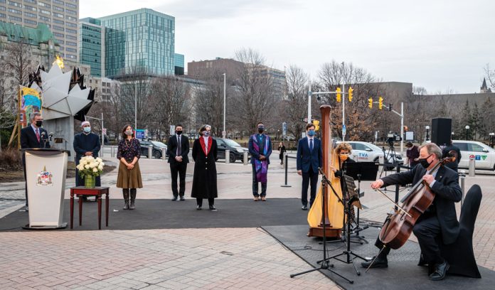 A group of people stand outside of city hall while two musicians play music on cellos on an overcast day in Ottawa|Mayor Jim Watson stands at a podium outside city hall on an overcast day in Ottawa with religious leaders standing behind him standing six feet apart and wearing masks