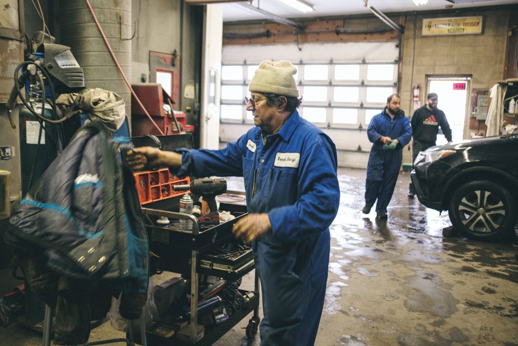 A man stands in a mechanic's uniform next to a red toolkit with cars and mechanics behind him