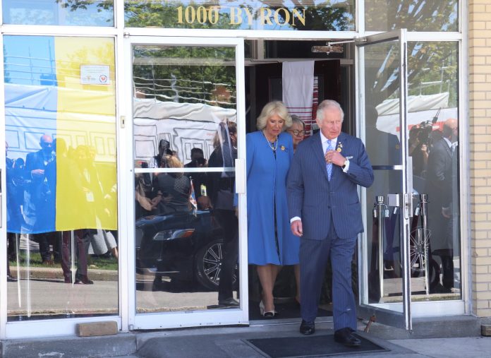 Prince Charles and Camilla walk out of the Ukrainian Orthodox Cathedral on Byron avenue. A Ukrainian flag hangs on the left.|A row of people stand outside the Ukrainian Cathedral hall waiting for the royals to leave the building.|A person wears a UK flag as a cape and walks across an open field on a sunny day.|Prince Charles wears a blue pinstripe suit and sunflower pin on a sunny day. There is a row of servicemen behind him.|Duchess Camilla is seen wearing a light blue suit with a sunflower pin and a pearl necklace. It is a sunny day and there is a royal guard in uniform behind her.