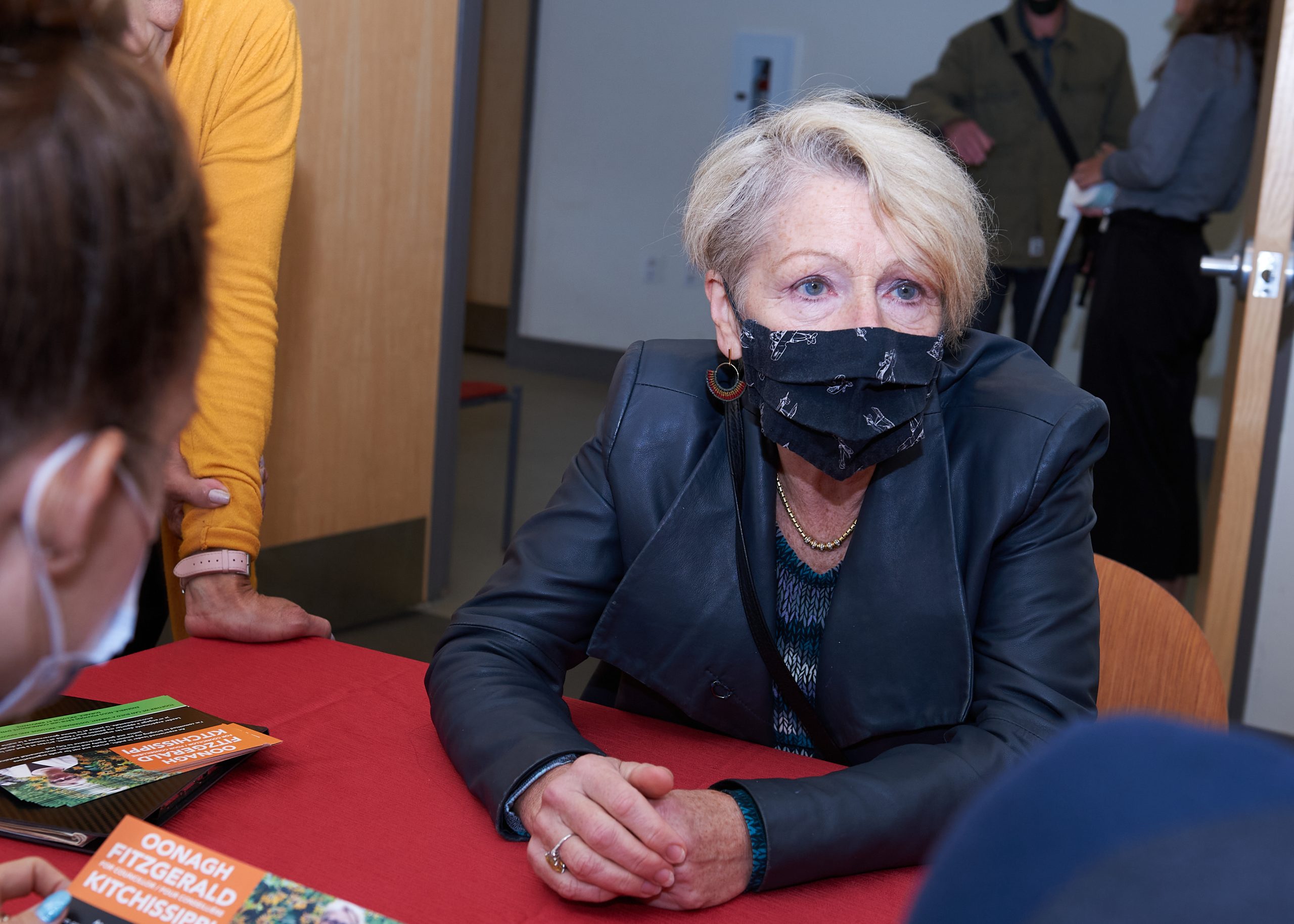 A senior woman wearing a black blazer, black face mask and black shirt sits at a table with her hands folded. There is a red tablecloth underneath her hands.