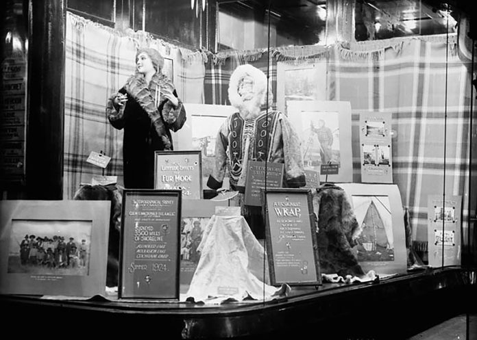 A black and white picture of a department store window featuring two manaquins wearing fur coats and several pictures in frames.||