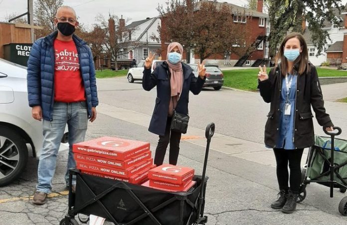 There people stand next to a black wagon carrying 15 red pizza boxes from Gabriel's Pizza on an overcast day in Ottawa|A grey cement wall with white graffiti is seen on a sunny day near Westboro Beach