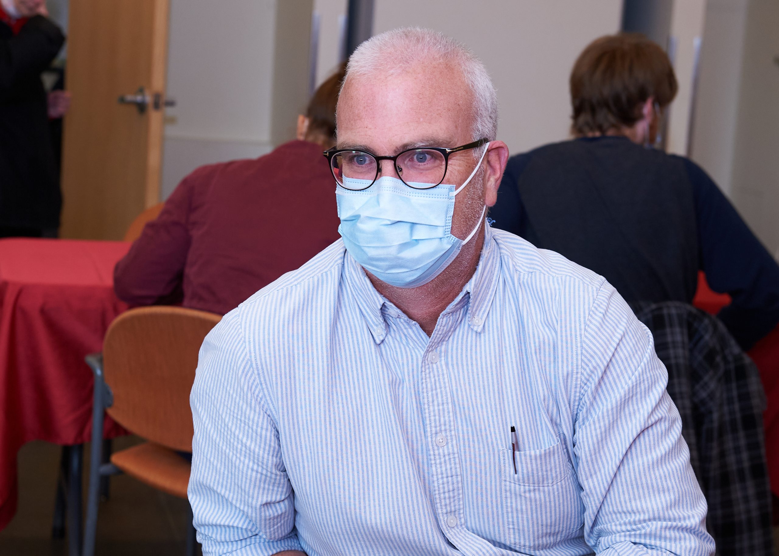 A man wearing a striped white shirt, blue face mask and black glasses sits at a table.