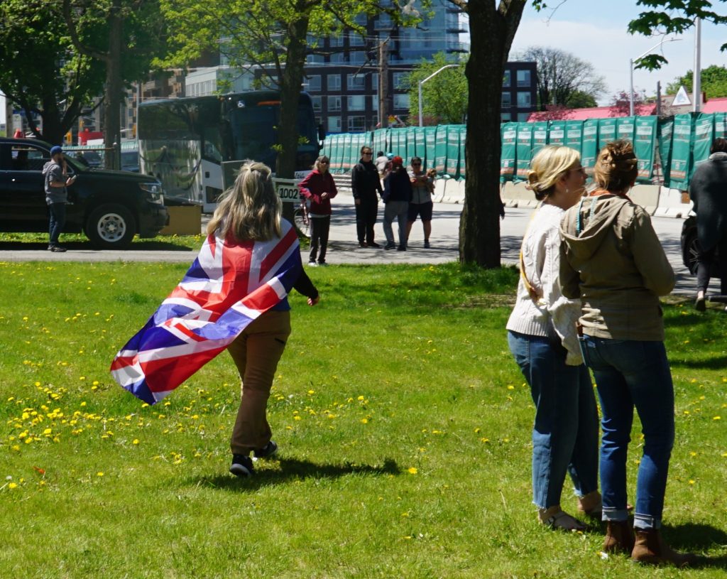 A person wears a UK flag as a cape and walks across an open field on a sunny day.