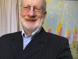 A headshot of Maciek Peter Kozlowski in front of a multicoloured abstract painting