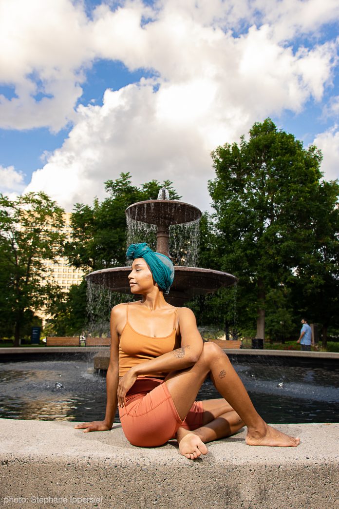 A Black woman holds a seated twist pose on a water fountain with a sunny sky behind her.|A Black woman holds a yoga pose outside of the National Art Gallery.|A Black woman holds a yoga pose on bended knee in between two pillars with a forest behind her.|An Asian woman holds a yoga pose with one arm outstretched standing on a beach