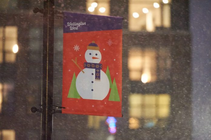 A snow covered sign by the Wellington West BIA with a snowman featured on it. The sign has a red and purple background.