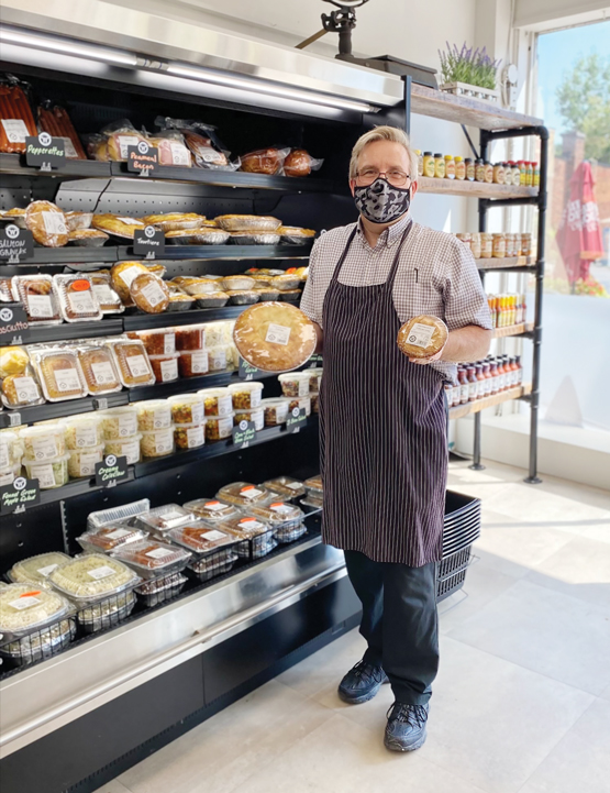 A man wearing a mask and butcher's apron stands in a story and holds up meat pies|