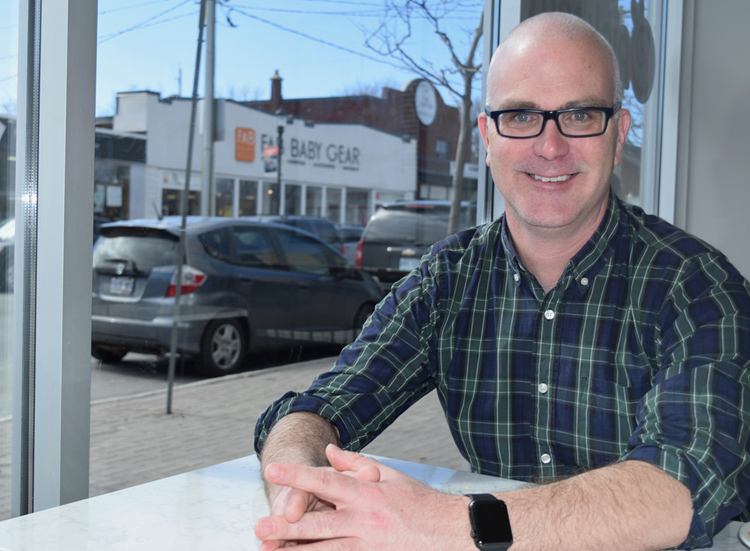 Kitchissippi Councillor Jeff Leiper is seen sitting at a table in Hintonburg. There is a sunny streetscape behind him.