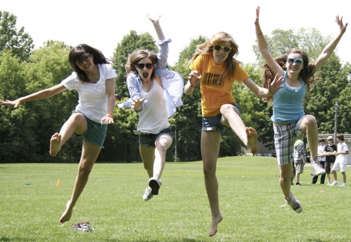 Four teenage girls jump in the air on a sunny day on a grass field.