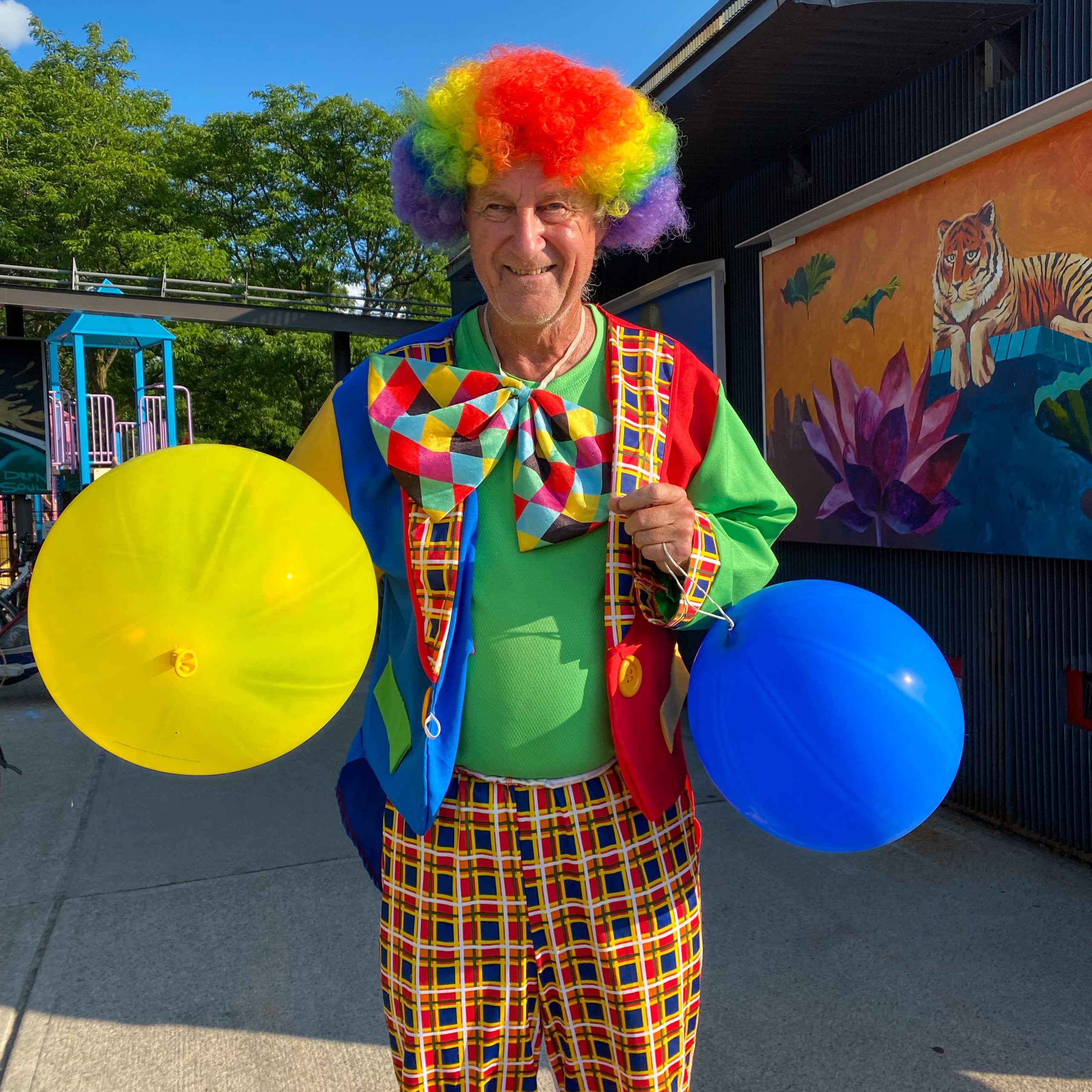 Busker the Balloon clown, wearing very colourful clothing, poses for a photograph at Parkdale Park. He’s holding balloons. 