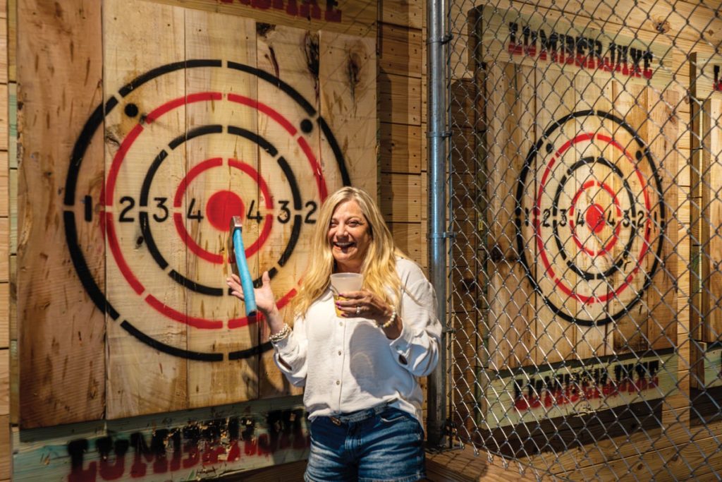 A woman with blonde hair holds a drink and an axe. 