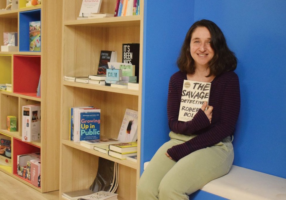 Shilhav sots on a bench and holds a book in her store. A blue wall is behind her and there is a bookshelf to the left. 