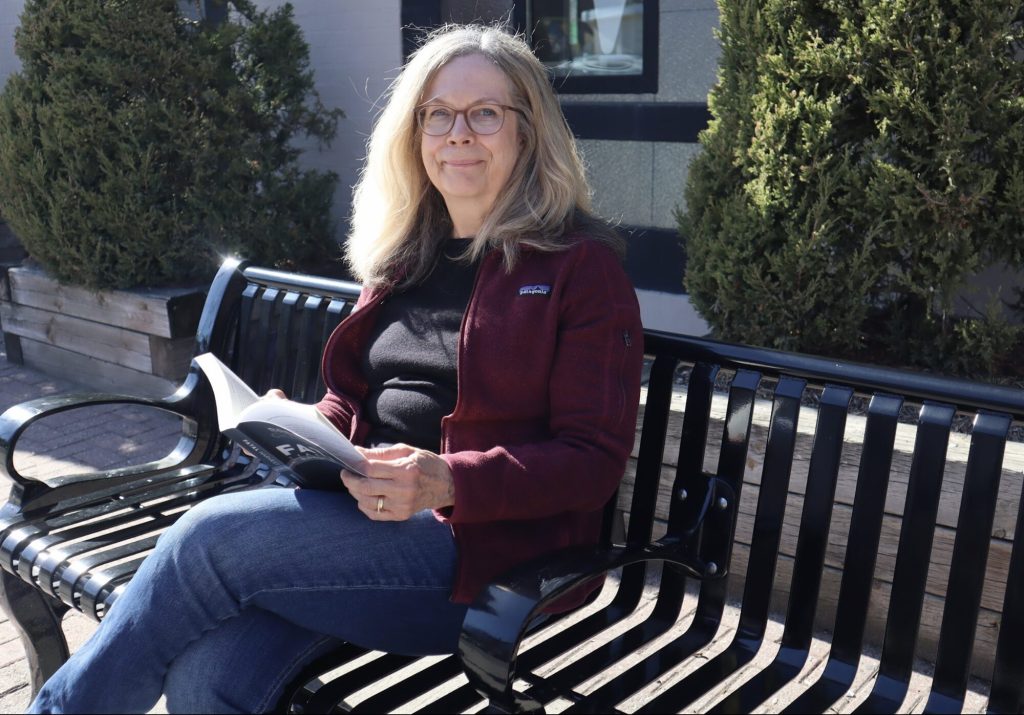 Brenda sits on a bench and holds up a copy of her book. 