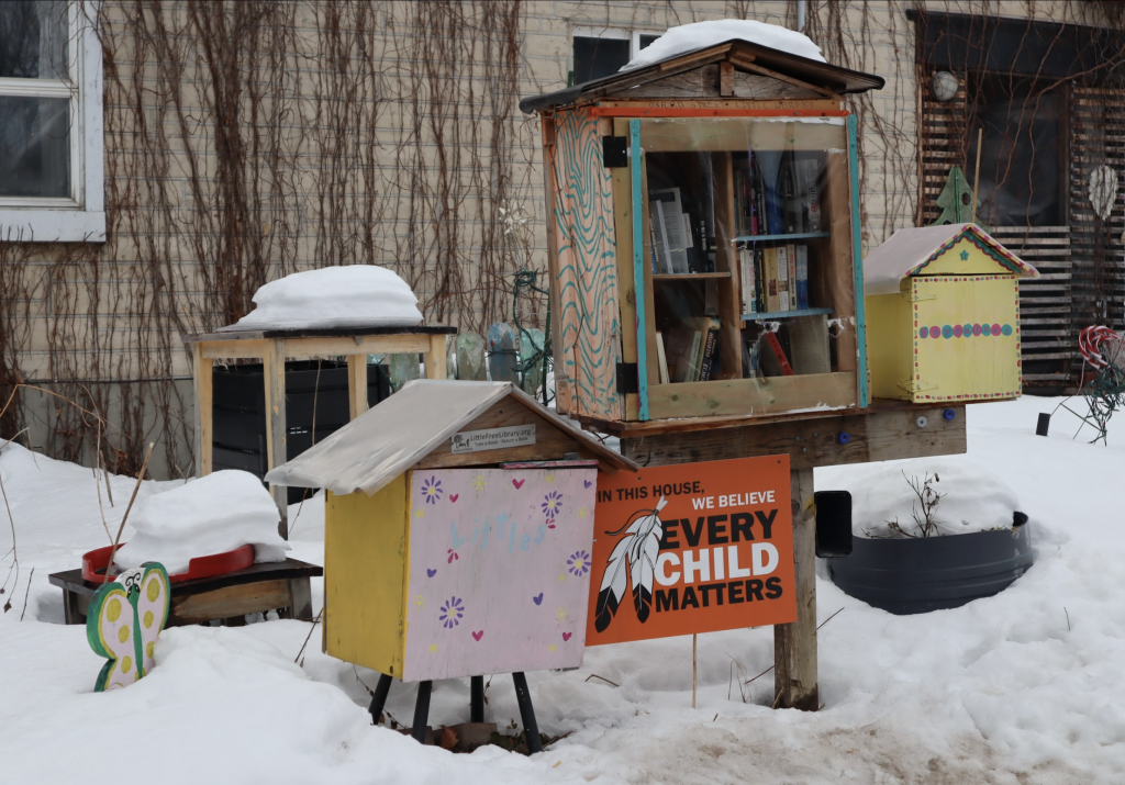 Three library boxes of different sizes sits outside a home. An orange sign reading “Every Child Matters” is also placed by the boxes.