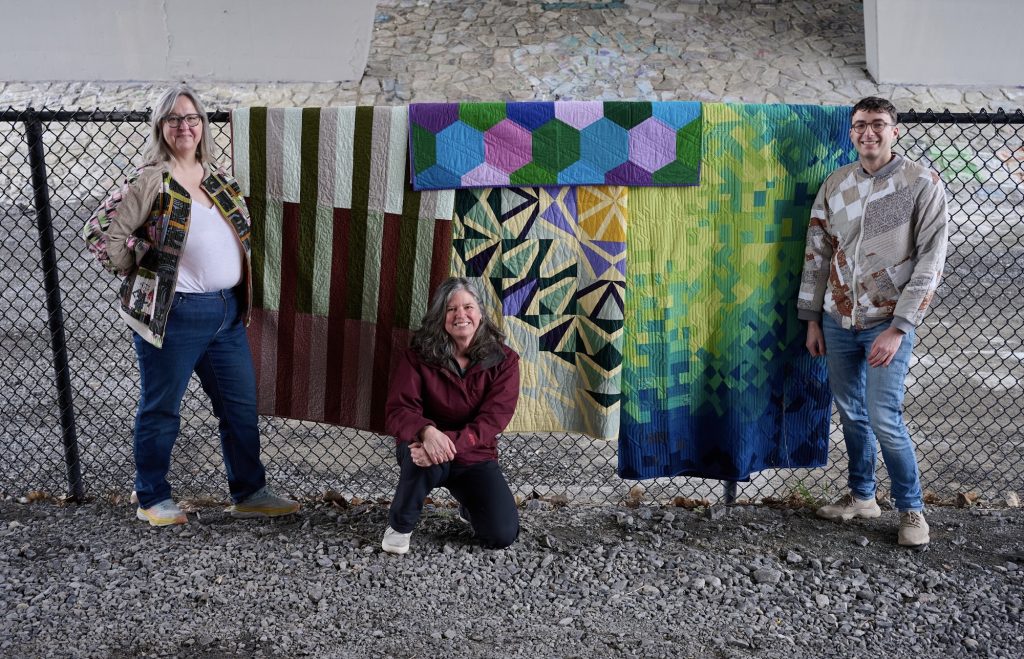 The three quilters pose for a photo with their quilts, which are hanging off a fence under a bridge. 