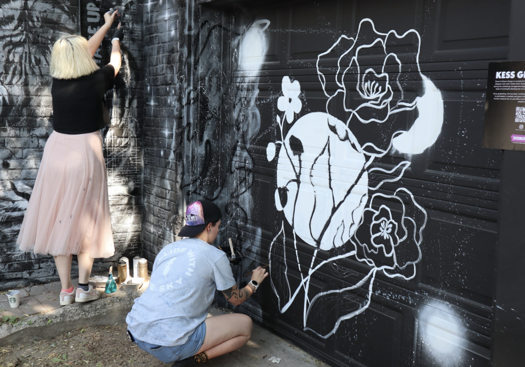 Two women paint murals on the side of Urban Art Collective. One design is of flowers. 