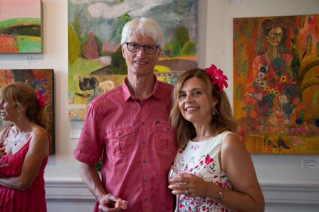 A man and a woman pose for a photograph with paintings behind them.