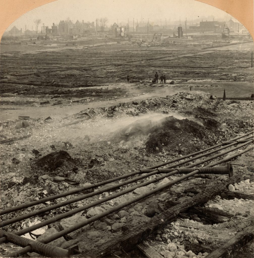 Burned remains of buildings are seen in the distance as a few people survey the damage. Streetcar tracks are noticeable at the front. 