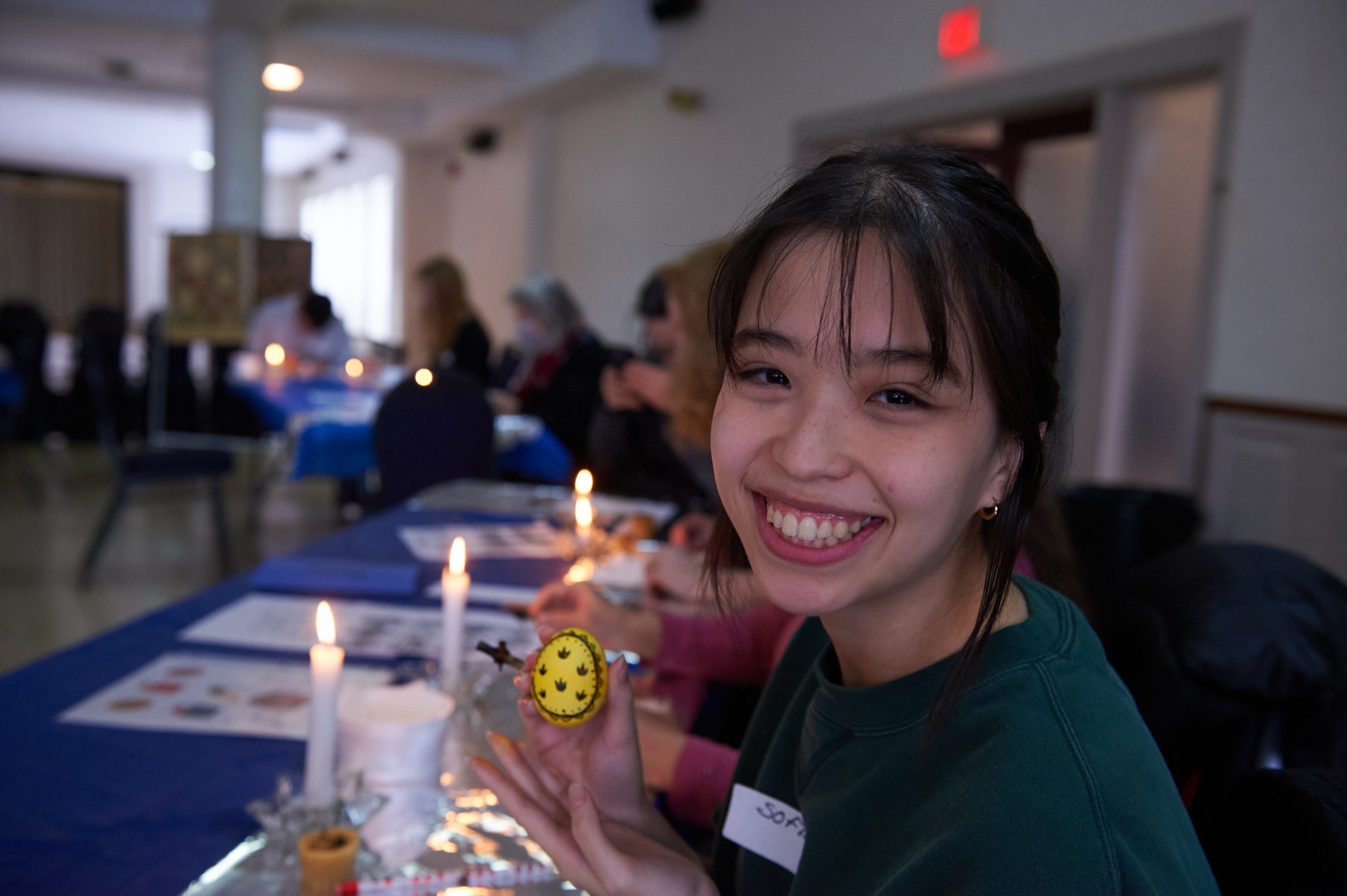 A woman with a big smile holds up a yellow painted egg with lines drawn on it.