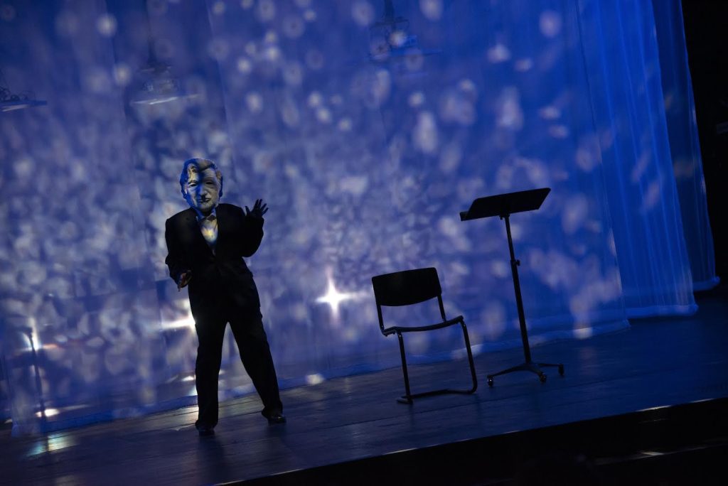 A person in a suit with a large face mould of a man is photographed on stage. The background has blue lights