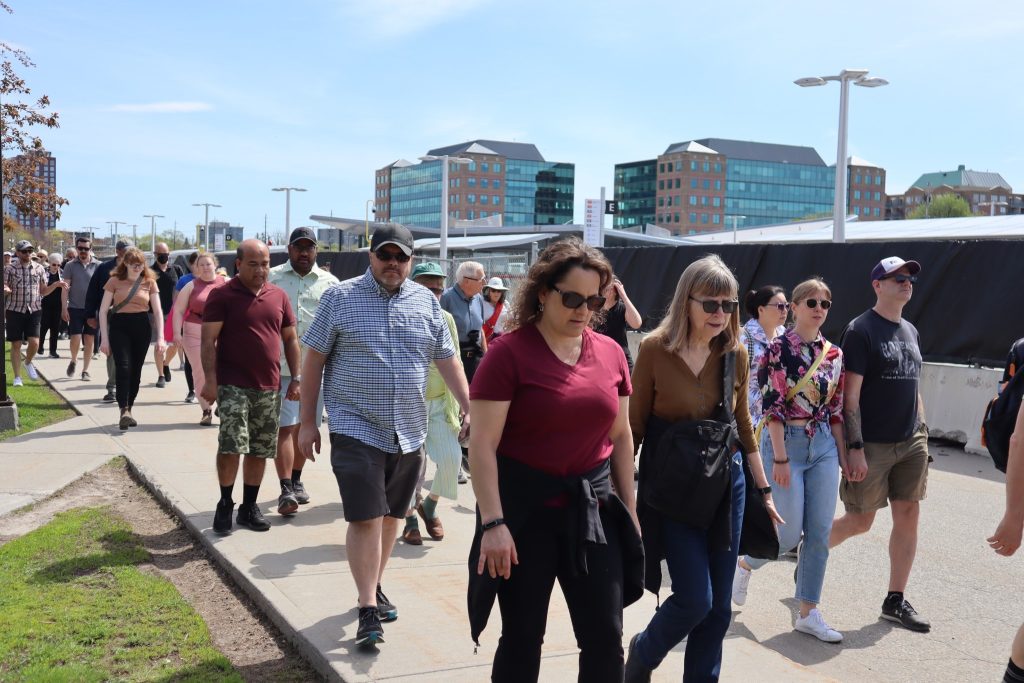 Many people walk down a sidewalk at Tunney’s Pasture. The bus station can be seen in the background.