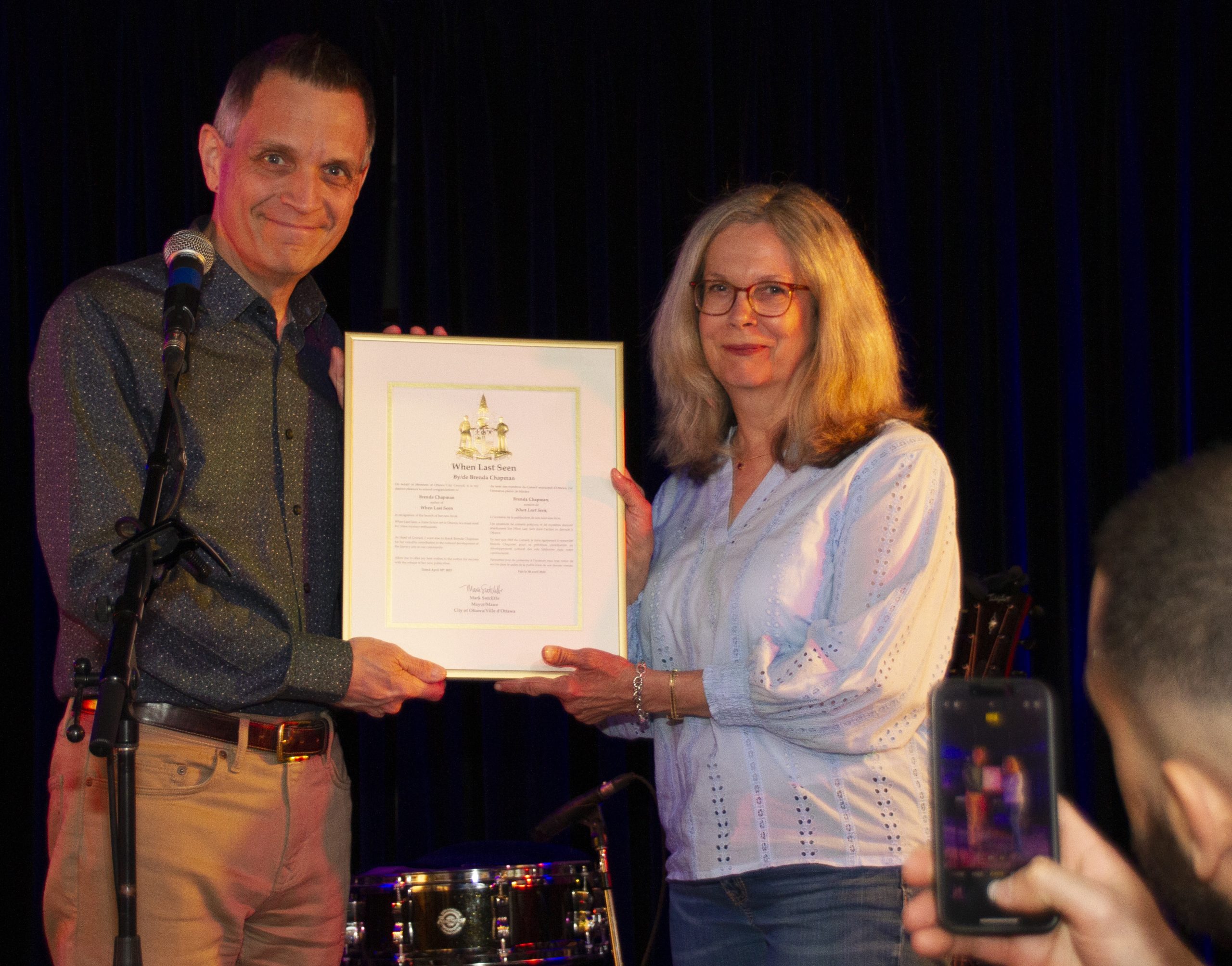 Author Brenda Chapman and Ottawa Mayor Mark Sutcliffe smile and pose for a photo as they hold up a plaque.