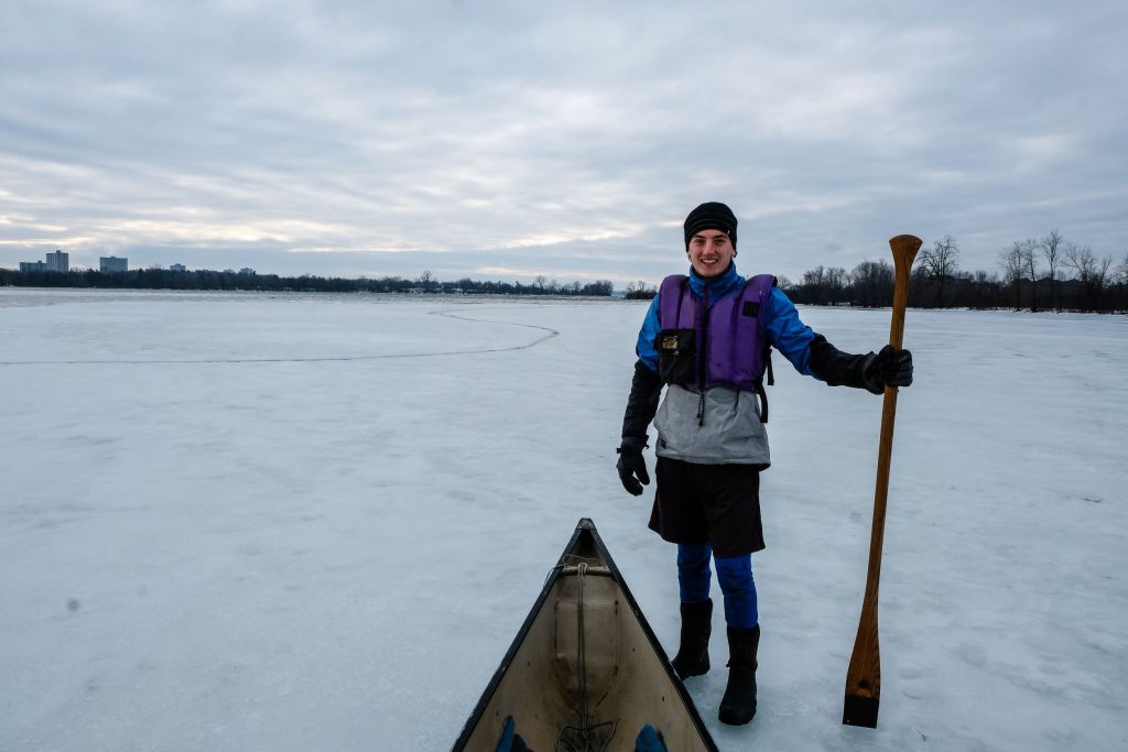 Isaac stands next to a canoe on the Ottawa River. It’s winter and the river is frozen over. 